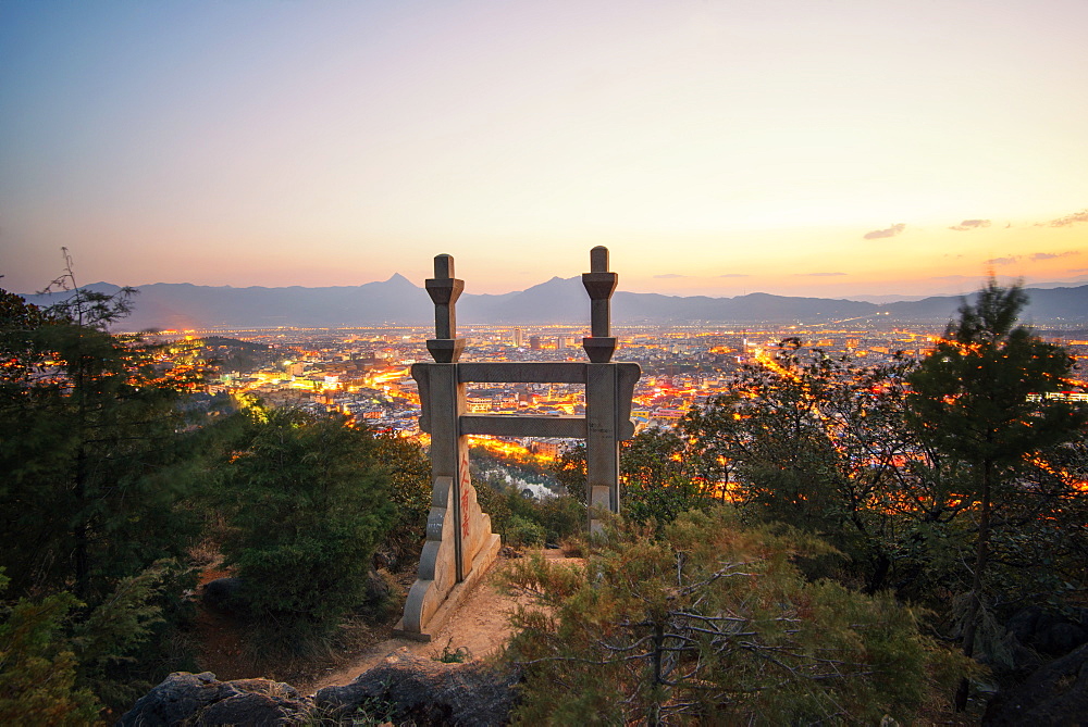 Stone gate in the mountains above Lijiang with foliage in the foreground and city lights in the background, Yunnan, China, Asia