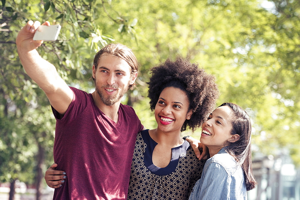 Three people, a man and two women, taking selfies in a city park