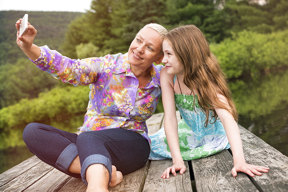 A woman and a child seated on a lake dock taking a selfie with a smart phone.