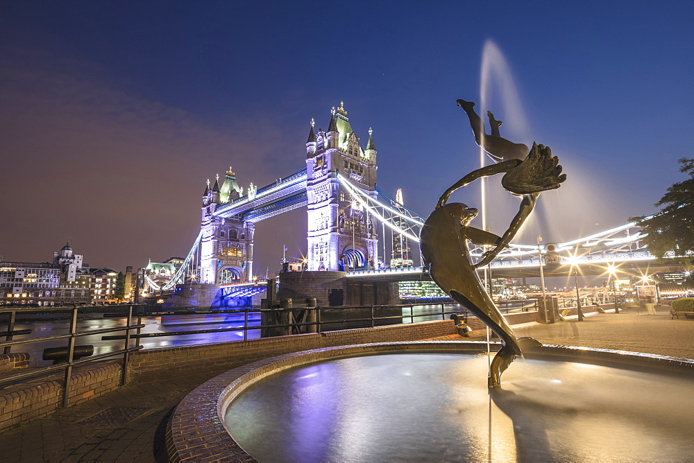 The Girl With A Dolphin Fountain frames Tower Bridge reflected in the River Thames at night, London, England, United Kingdom, Europe