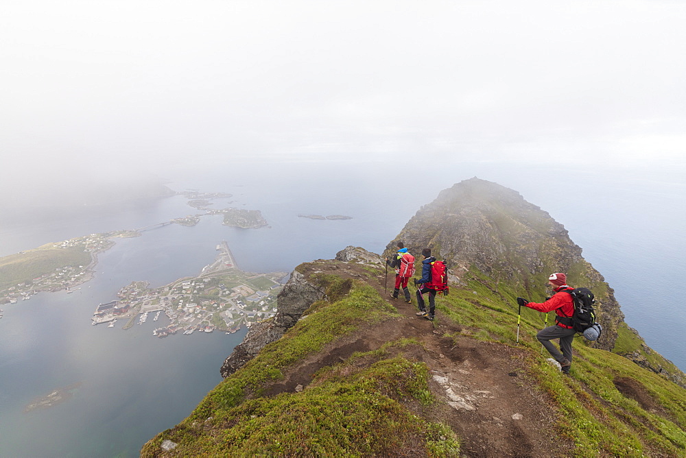 Hikers on top of rocky peak admire the blue sea surrounded by mist, Reinebringen, Moskenesoya, Lofoten Islands, Norway, Scandinavia, Europe