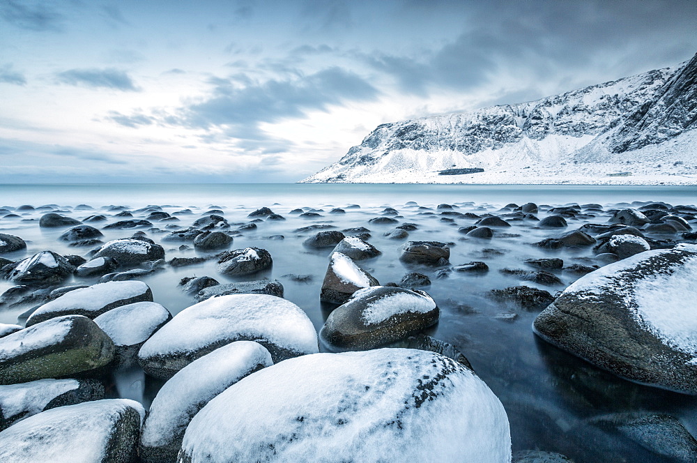 Rocks in the cold sea and snow capped mountains under the blue light of dusk, Unstad, Lofoten Islands, Arctic, Northern Norway, Scandinavia, Europe