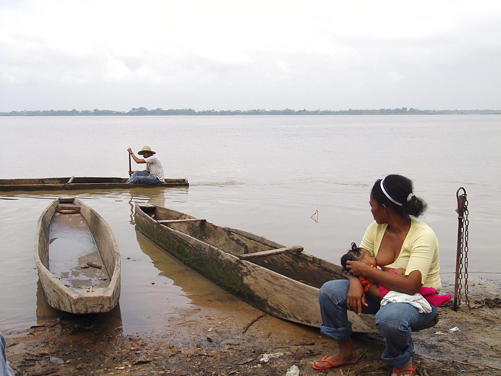 Colombia woman breast feeding her baby beside the rio magdalena, barrancabermeja. Fishermen go passed in a dug our canoe