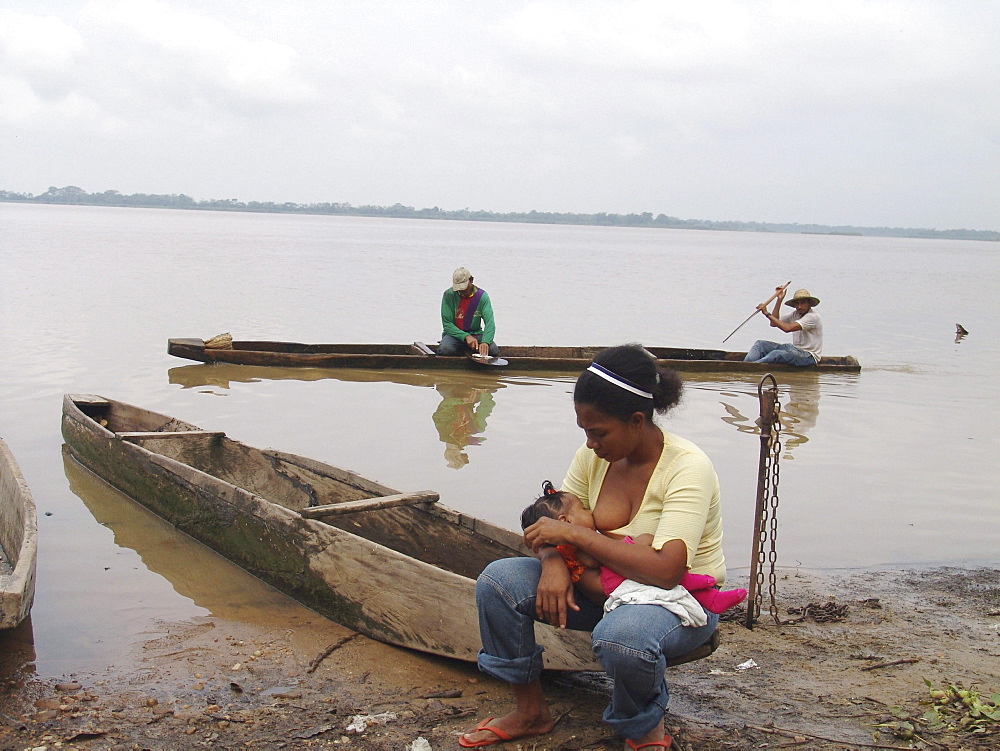 Colombia woman breast feeding her baby beside the rio magdalena, barrancabermeja. Fishermen go passed in a dug our canoe