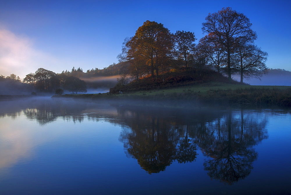 A calm clear autumn morning at Elterwater as the first light of a new day illuminates the trees and mist, Lake District National Park, UNESCO World Heritage Site, Cumbria, England, United Kingdom, Europe