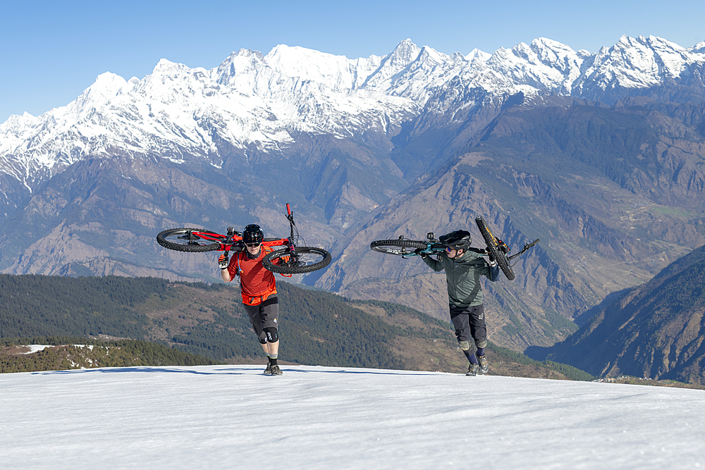 Mountain bikers carry their bikes up a snow covered slope in the Himalayas with views of the Langtang range in the distance, Nepal, Asia