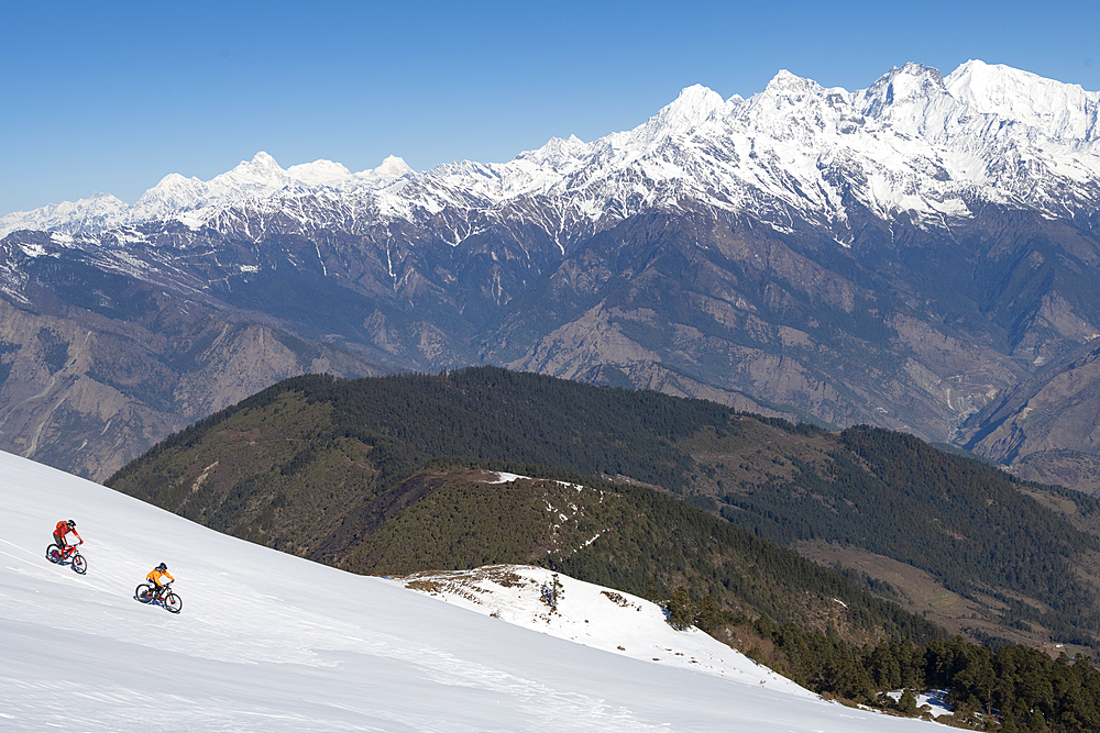 Mountain bikers cycle along a snow covered slope in the Himalayas with views of the Langtang range in the distance, Nepal, Asia