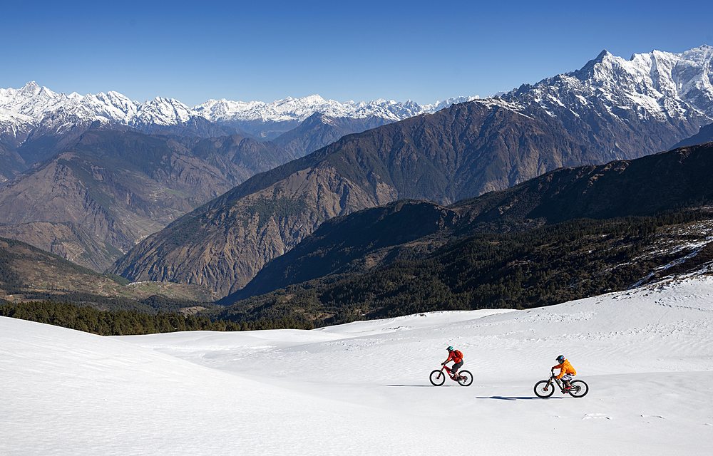 Mountain bikers descend a snow covered slope in the Himalayas with views of the Langtang range in the distance, Nepal, Asia