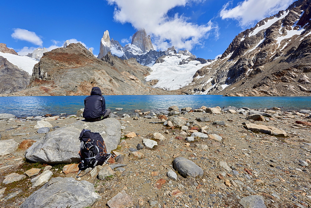 A lone figure in mountain gear rests on rocks with view to Lago de los Tres and Mount Fitz Roy, their backpack on the ground, Patagonia, Argentina, South America