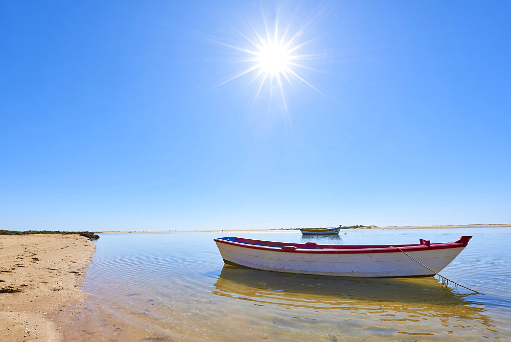 The sun shines above a small fishing boat on transparent lagoon water in Cacela Velha, Algarve, Portugal, Europe