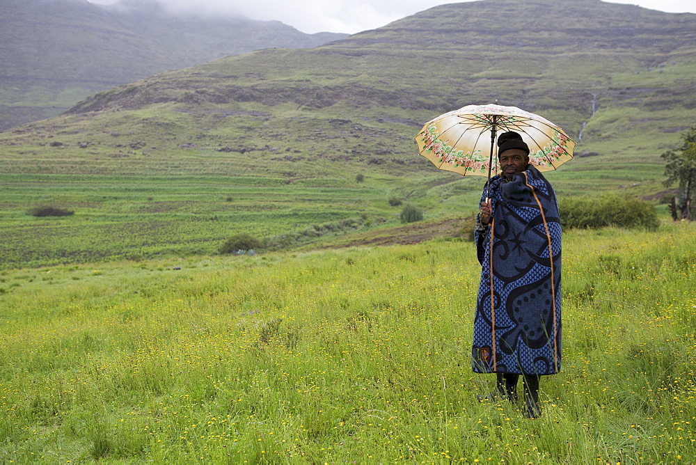 A man holding an umbrella and wrapped in a blue blanket stands in the mountains, Lesotho, Africa