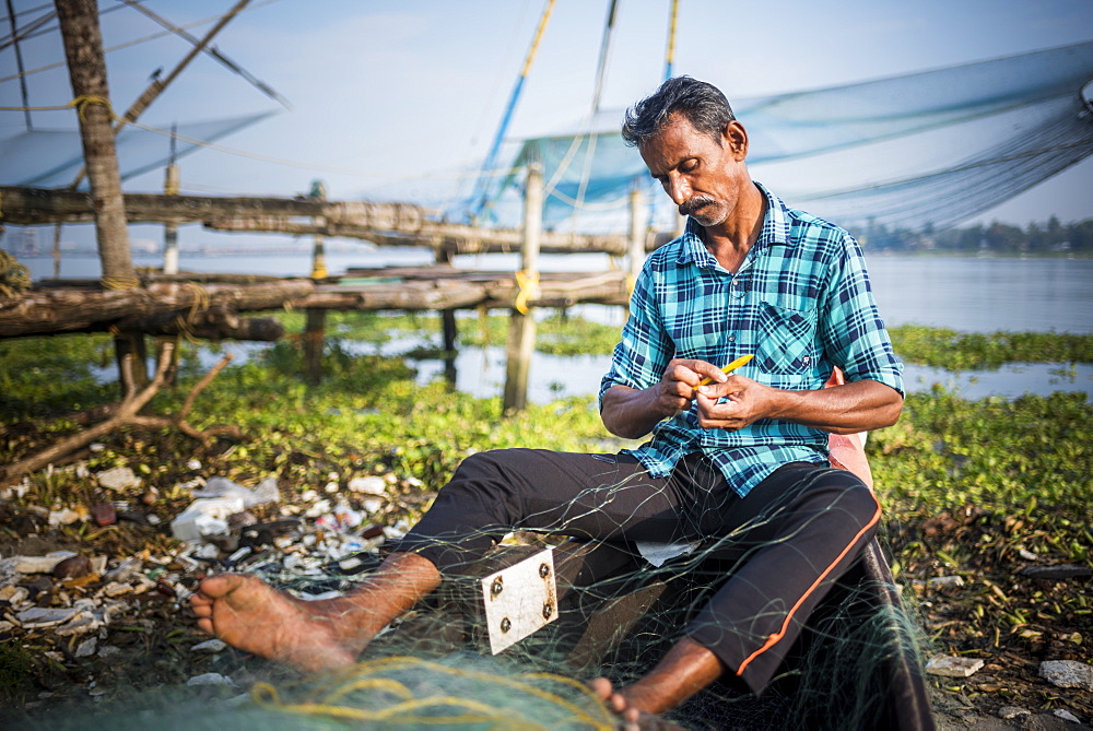 Fisherman mending net with traditional Chinese fishing nets behind, Fort Kochi (Cochin), Kerala, India, Asia