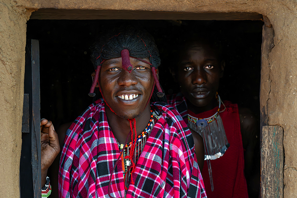 Two Maasai tribesmen in costume wearing traditional dress stood in doorway of mud house, Maasai Mara, Kenya Africa