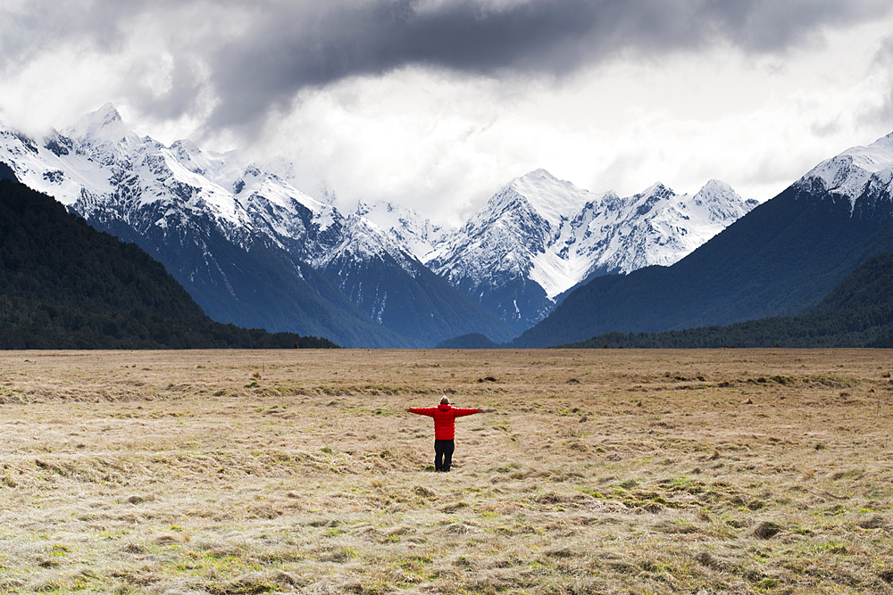 Man in red coat stood holding out his arms looking at snow capped mountains, Fiordland National Park, UNESCO World Heritage Site, South Island, New Zealand, Pacific