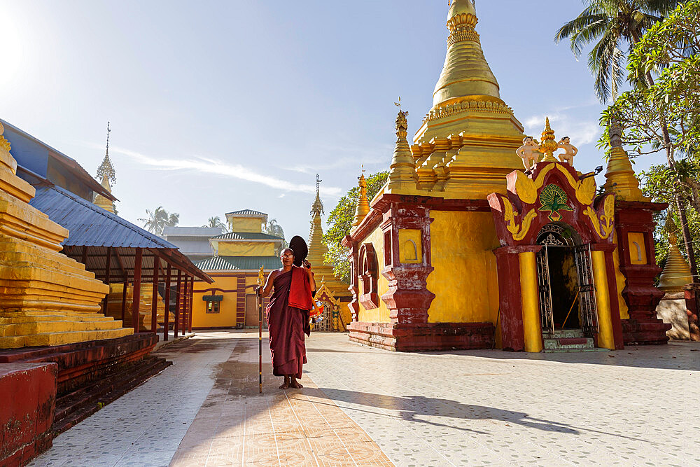 A monk in maroon robes holding a staff and a steel alms bowl standing in the grounds of a temple, Sittwe, Rakhine, Myanmar (Burma), Asia