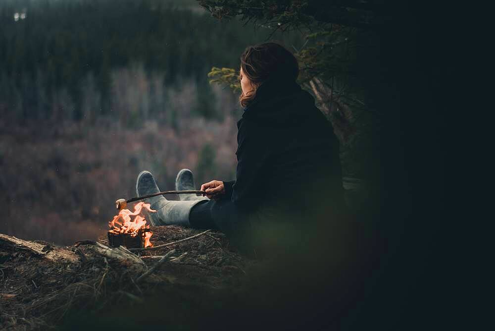 Woman roasting a marshmallow while camping along the edge of a cliff. Yukon Territory, Canada