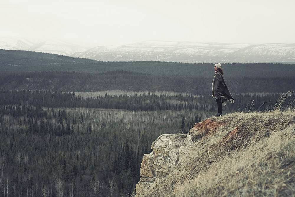 A young Woman is standing at the edge of a cliff with an endless view over forest and mountains. Yukon Territory, Canada