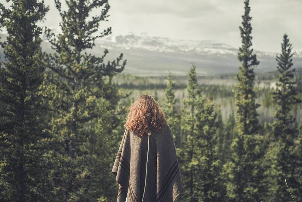 A young woman enjoys the view over forest and mountains. Yukon Territory, Canada