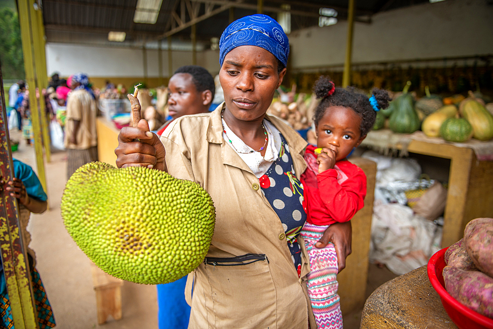 A woman holds her child under her arm while examining a jackfruit at outdoor market, Rwanda Farmers Market, in Rwanda