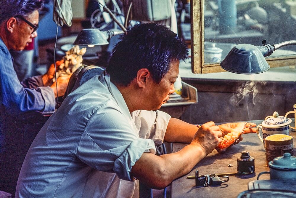 An artisan in a jade factory works on a jade figurine whlie smoking a cigarette. Beijing, China.