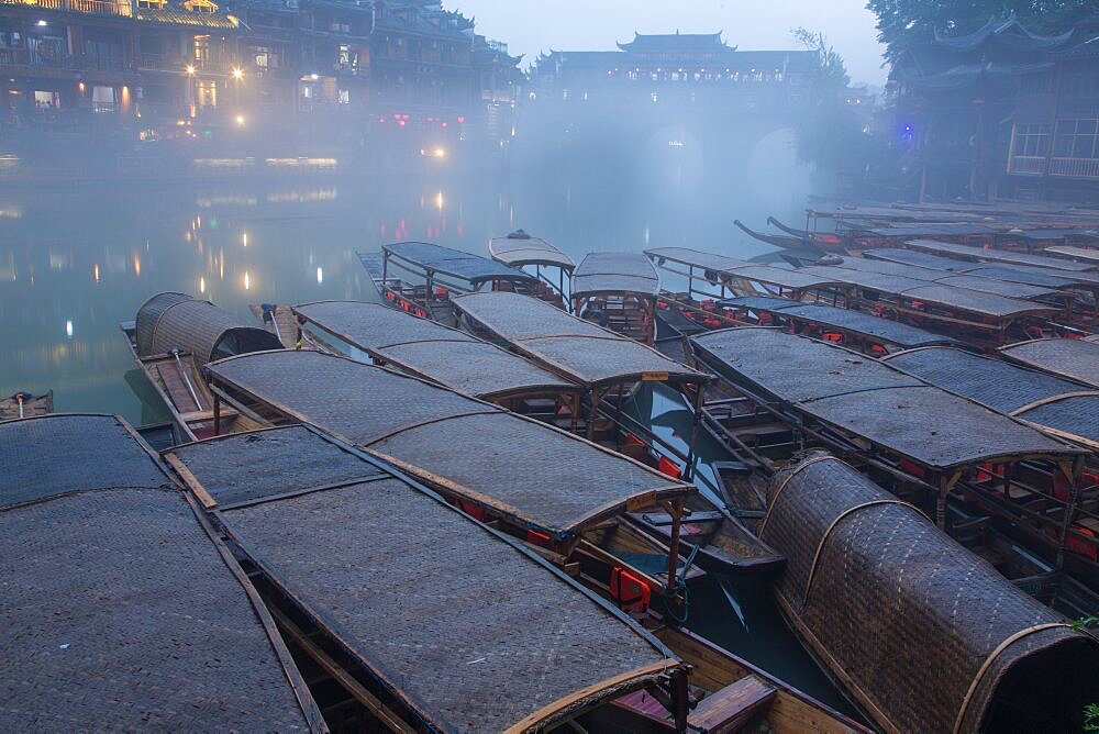 Covered tour boats docked on the bank of the Tuojiang River. The Phoenix Hong Bridge is in fog. Fenghuang, China.