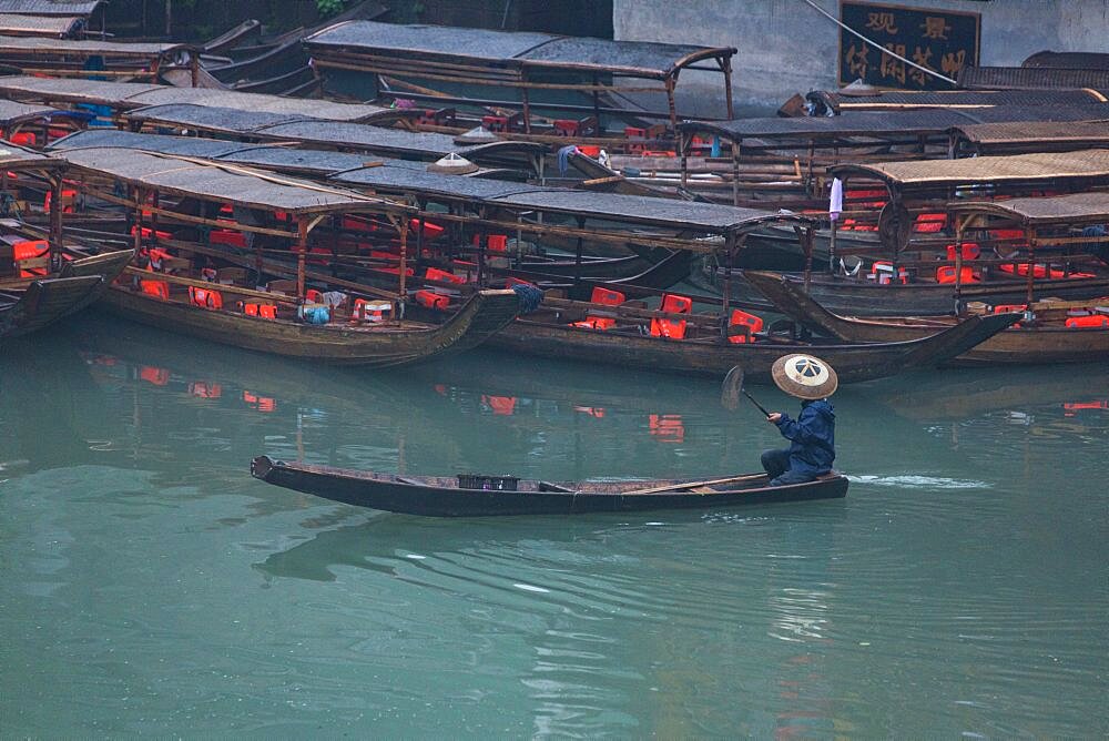 A man in a traditional conical hat paddles a sampan on the Tuojiang River in Fenghuang, China.