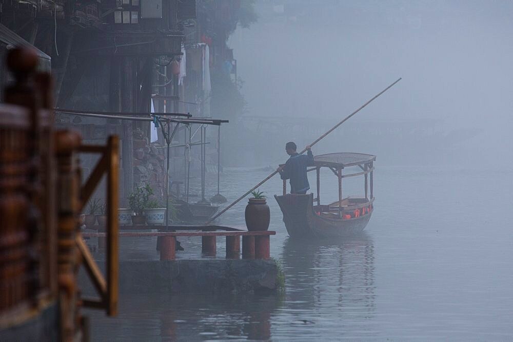 A man poles an empty covered tour boat up the Tuojiang River in early-morning fog. Fenghuang, China.