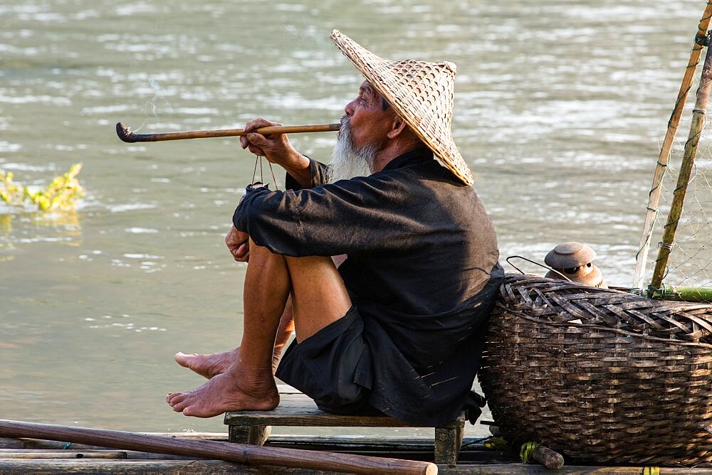 Elderly traditional cormorant fisherman smokes a wooden pipe on his raft on the Li River, Xingping, China.