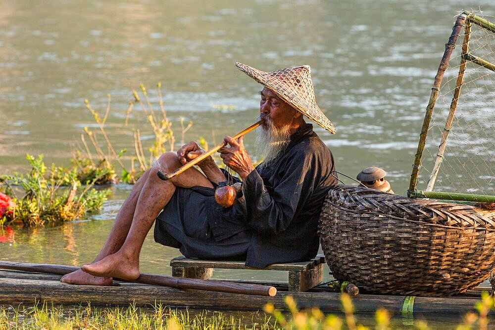 Elderly traditional cormorant fisherman smokes a wooden pipe on his raft on the Li River, Xingping, China.