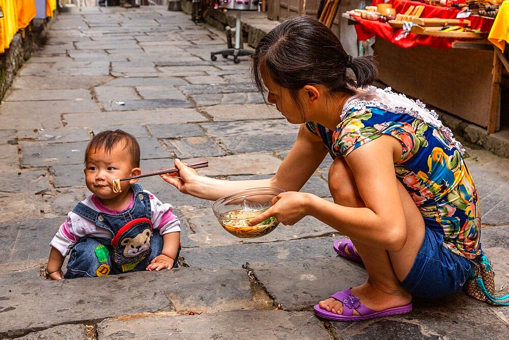 A young Chinese mother feeds her baby boy noodles with chopsticks on a cobblestone street. Furong, China.