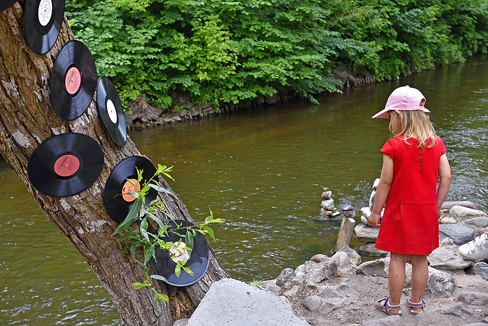 Little girl in a red dress on the Vilnia River bank next to tree decorated with vinyl records, Uzupis district, Vilnius, Lithuania, Europe