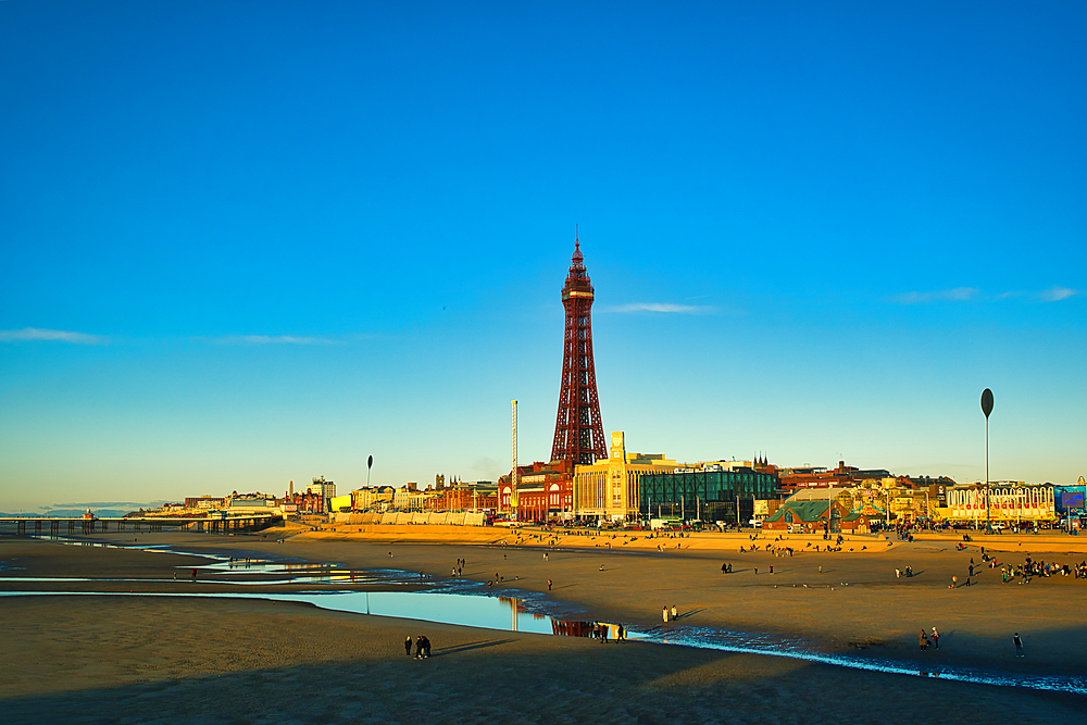 Golden hour view of a seaside town with a prominent tower, clear blue sky, and reflections on the wet sand in Blackpool, England