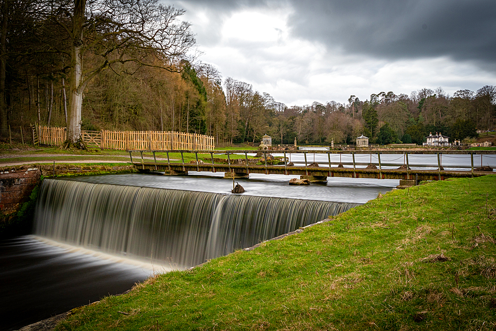 Tranquil river waterfall with smooth flowing water, surrounded by lush trees and overcast skies, United Kingdom, Europe