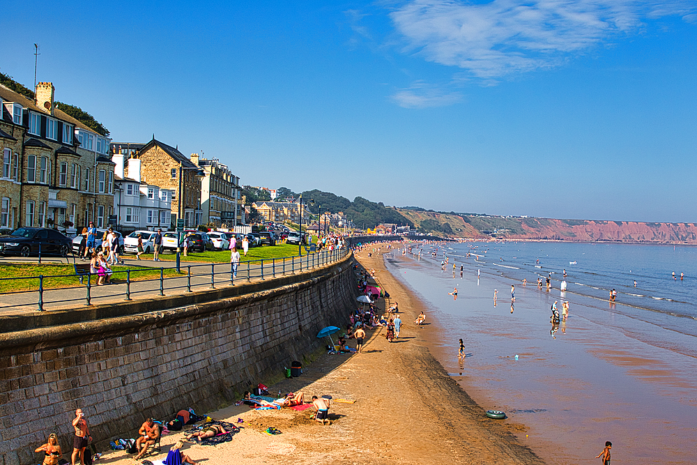 Sunny beach day with people relaxing and walking along the shore, coastal buildings in the background in Filey, England.
