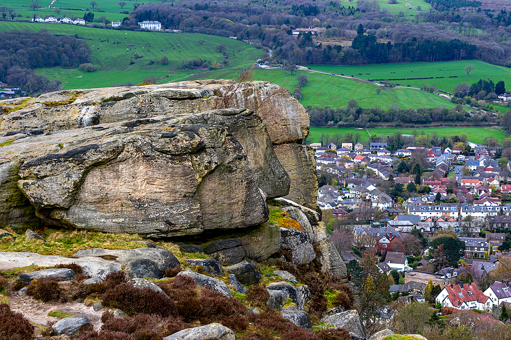 Scenic view of a large rock formation overlooking a lush valley with a small town and green fields in the background in Ilkley, North Yorkshire, England, United Kingdom Europe