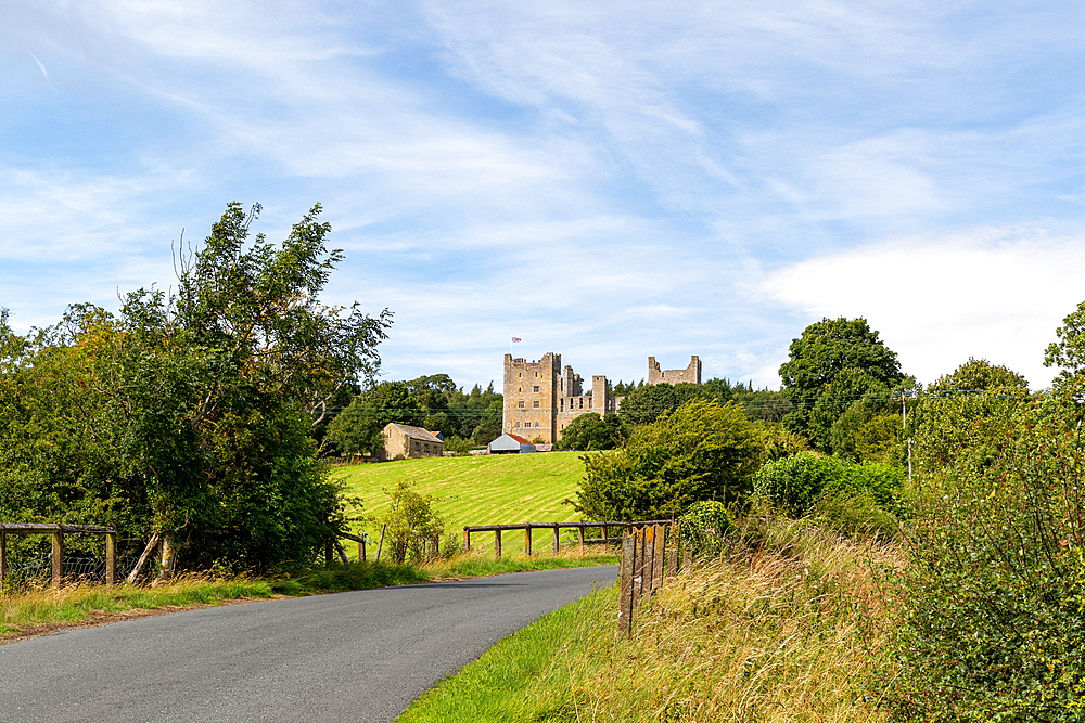 Rural road leading to an ancient castle amidst green fields under a blue sky with clouds in Yorkshire Dales, Yorkshire, England, United Kingdom, Europe