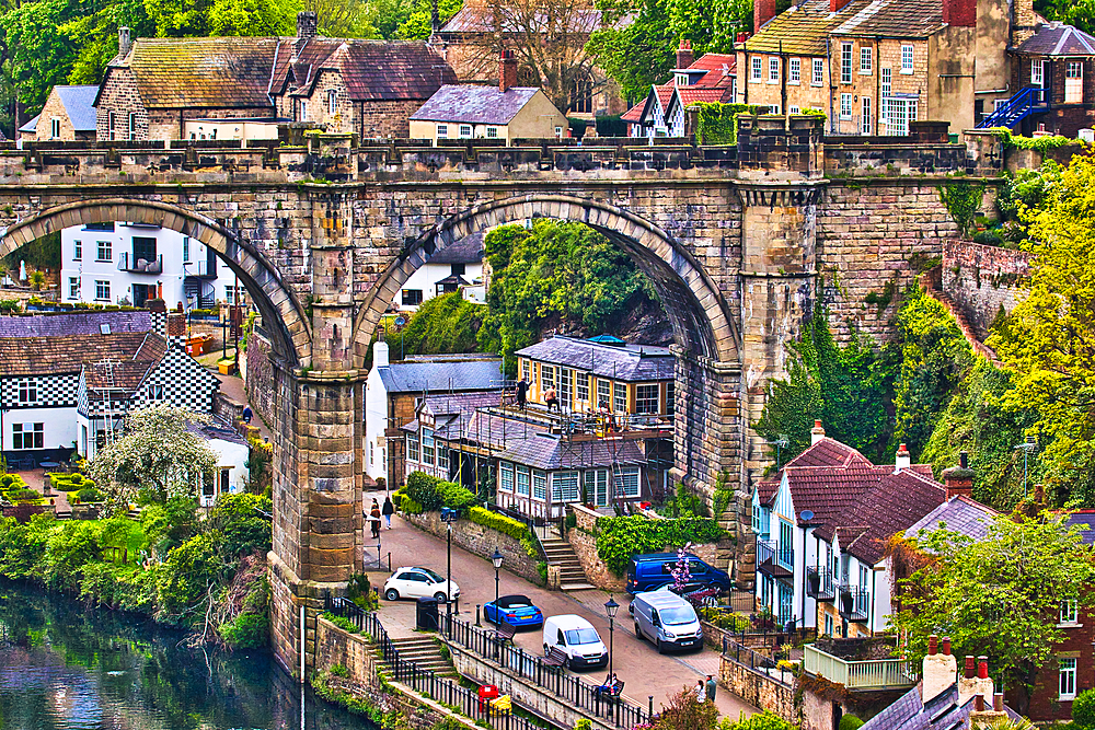 A picturesque view of a quaint village with an old stone bridge arching over a river, surrounded by lush greenery and traditional houses.