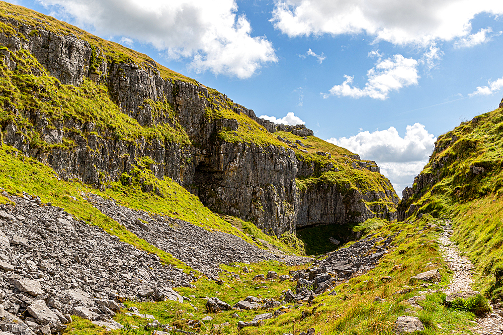 Lush green landscape with rocky cliffs and a clear blue sky, featuring a natural cave entrance and footpath at Malham Cove, Yorkshire, England, United Kingdom, Europe