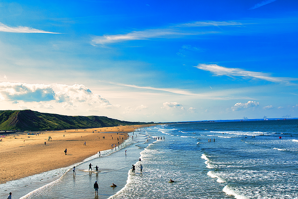 Sunny beach scene with people and rolling waves, blue sky with clouds overhead, and a hilly backdrop in Saltburn-by-the-Sea, England.