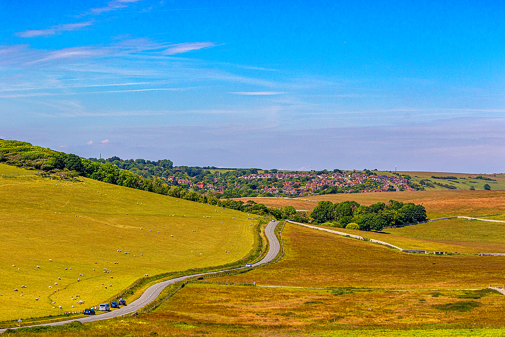 Scenic countryside road winding through lush green fields under a clear blue sky looking towards East Dean, South Downs National Park, East Sussex, England, United Kingdom, Europe