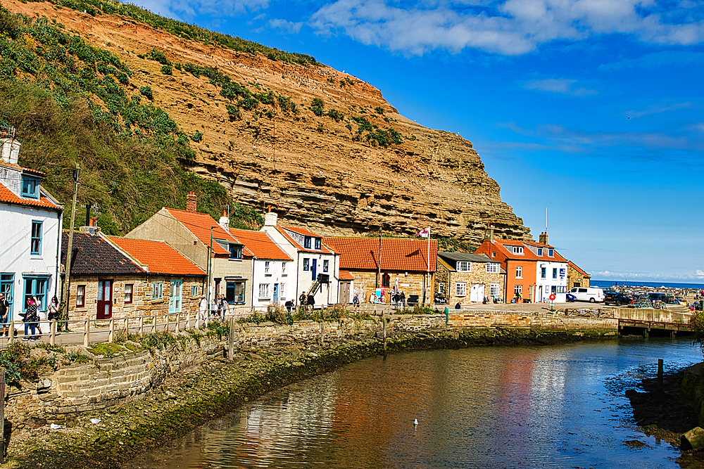 Quaint coastal village with traditional houses along a river, under a clear blue sky with a rocky cliff in the background in Staithes, England.