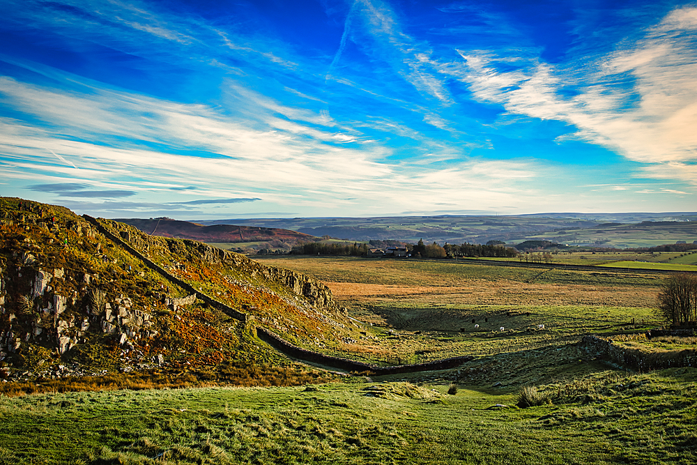 Scenic view of a lush countryside with a stone wall under a blue sky with wispy clouds at Sycamore Gap, Northumberland, UK.