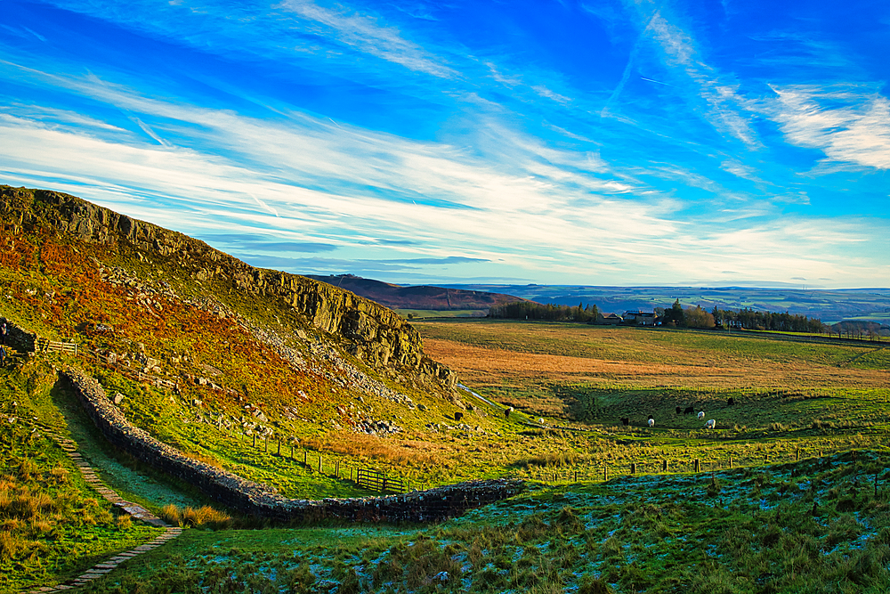 Scenic landscape with vibrant blue skies, wispy clouds, and a rocky hillside overlooking a lush green valley at Sycamore Gap, Northumberland, UK.