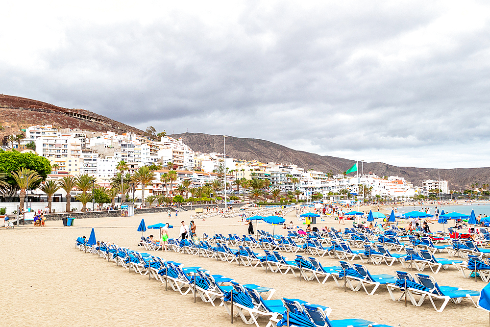 Crowded beach with rows of blue sun loungers, white buildings in the background under a cloudy sky in Los Cristianos, Tenerife, Canary Islands, Spain, Atlantic, Europe