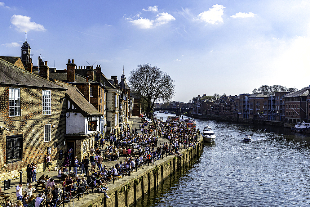 Scenic riverside with people enjoying sunny weather at outdoor cafes and boats on the water, against backdrop of historic buildings in York, North Yorkshire, United Kingdom, Europe