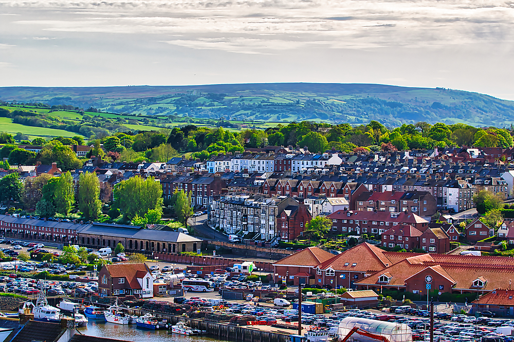 Aerial view of a coastal town with a harbor filled with boats. The town features a mix of residential buildings and greenery, with rolling hills in the background under a partly cloudy sky.