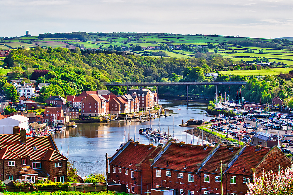 A scenic view of a small town with red-roofed houses by a river, a bridge crossing the river, and lush green hills in the background. The town has a marina with several boats docked.