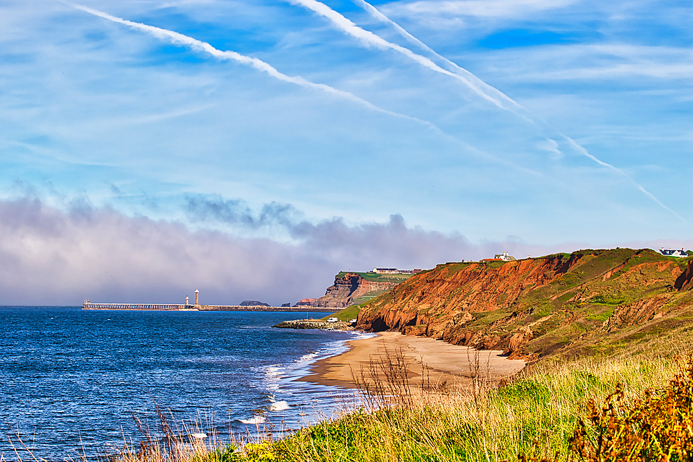 A scenic coastal landscape featuring a sandy beach, cliffs, and a distant pier with lighthouses. The sky is blue with some clouds and contrails.