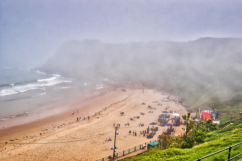 A foggy beach scene with people relaxing on the sand and a few tents set up. The ocean waves are gently crashing onto the shore, and the background is obscured by dense fog.