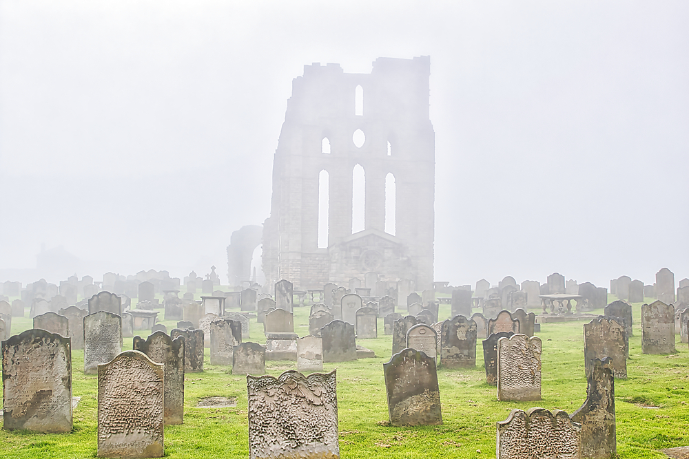 A foggy cemetery with old tombstones and a large, ancient, ruined building in the background.